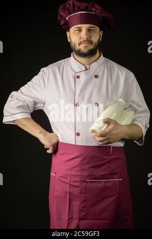 Gutaussehender bärtiger Chefkoch in Uniform hält zwei Flaschen mit Milch Stockfoto