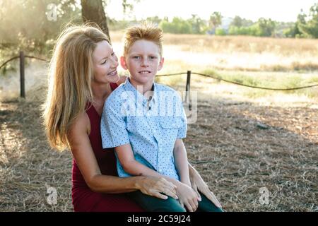 Ein süßer Junge mit rotem Haar sitzt draußen auf der Schote seiner Mutter Stockfoto