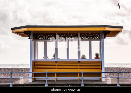 Ältere Menschen saßen an einem regnerischen Tag in Southsea, Portsmouth, Großbritannien, auf einer Bank unter einem viktorianischen Schutzhaus am Meer Stockfoto