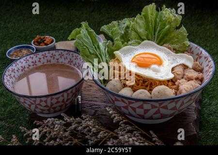 Eiernudeln mit Schweinefleischkugel, Hackfleisch, Spiegelei, Gemüse und Suppen in Schüssel und Essstäbchen mit Löffel und Holz Hintergrund kochen. Thailändische Nudelsuppe Stockfoto