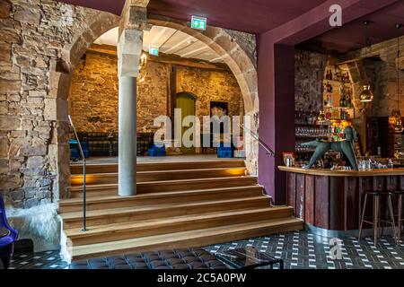 Ein historischer Steinbogen mit freiliegendem Schuttmauerwerk umrahmt eine Holztreppe, die in die Lounge des Hotel Renthof in Kassel, Deutschland, führt – das ehemalige Karmeliterkloster, gegründet 1298, das im Juni 2017 als Hotel, Restaurant und Veranstaltungsort wiedereröffnet wurde, zeitgleich mit documenta 14. "Jeder Bogen ist ein Geschenk," sagt Miteigentümerin Kirstin Homburg-Kleinkauf über den Bogen, der den Eingangsbereich mit dem Bistro und der Bar verbindet. Laut einer Beobachtung des Hauses neigen die Augen der Männer beim Betreten der Bar dazu, sich auf die Statik des alten Steins zu legen, während der Blick der Frauen zu den samtigen Armchais wandert Stockfoto