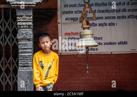 Ein kleiner Junge blickt auf eine Votivglocke, die in der Lobby eines nepalesischen buddhistischen Tempels in Thimi, Kathmandu Valley, Nepal, hängt. Stockfoto