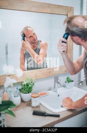 Lächelnder Mann, der neue Art Haarschnitt ein Haar mit einem elektrischen wiederaufladbaren Trimmer suchen im Badezimmerspiegel. Frisur, Körper und Hautpflege Stockfoto