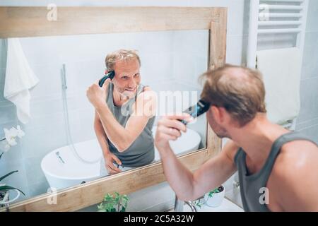 Lächelnder Mann, der neue Art Haarschnitt ein Haar mit einem elektrischen wiederaufladbaren Trimmer suchen im Badezimmerspiegel. Frisur, Körper und Hautpflege Stockfoto