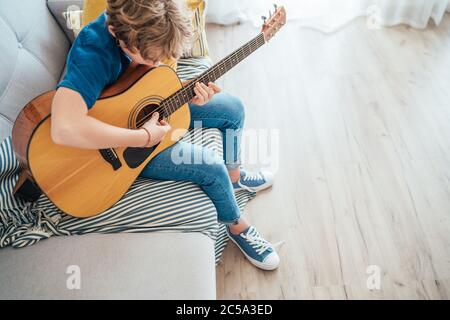 Pre teen Junge spielen akustische Gitarre gekleidet lässige Jeans, Hemd und neue Sneakers sitzen auf dem gemütlichen Sofa zu Hause Wohnzimmer. Musikerziehung top ang Stockfoto