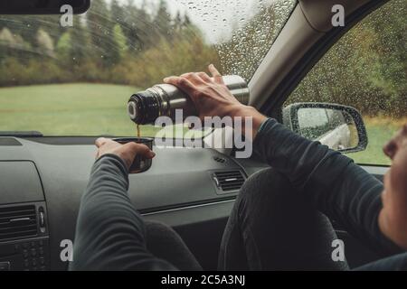 Frau gießt den heißen Tee in Touristen Thermoskanne. Sie sitzt auf dem Beifahrersitz im modernen Auto und genießt das launische regnerische Wetter, das durch die Nacht schaut Stockfoto
