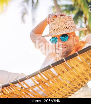 Junger Mann in Strohhut und blauer Sonnenbrille liegt schwingend in gemütlichen Hängematte aufrichtig lächelnd treffen Morgensonne Sonnenlicht am Strand unter der Palme Stockfoto