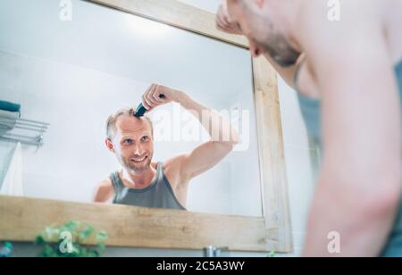 Lächelnder Mann, der neue Art Haarschnitt ein Haar mit einem elektrischen wiederaufladbaren Trimmer suchen im Badezimmerspiegel. Neue Frisur, Körper und skinc Stockfoto