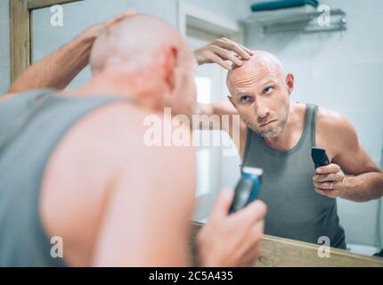 Glatze Mann berühren seinen neuen Stil Glatze Kopf Frisur machte er mit einem elektrischen wiederaufladbaren Trimmer suchen im Badezimmer Spiegel. Radikale Entscheidung, Life-ch Stockfoto