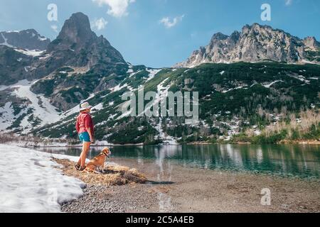 Junge Hündin bekleidet ein hellrotes Hemd, die mit seinem Freund Beagle an der Bergseenküste entlang wandern.Sie genießen Frühlingsstimmung und schmelzenden Schnee Stockfoto