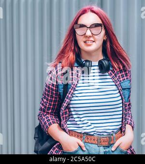 Schöne moderne lächelnde junge Teenager mit außergewöhnlicher Frisur Farbe in einem karierten Hemd und Spiegelsonnenbrille Porträt mit Kopfhörer Stockfoto