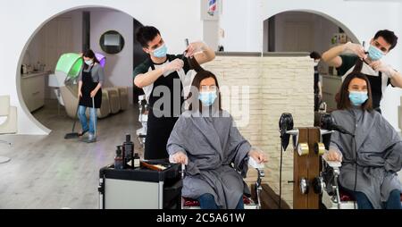 Positive männliche Friseur in Schutzmaske und Handschuhe machen Haar Styling für junge Frau, Arbeitstag im Schönheitssalon während Pandemie-Situation Stockfoto