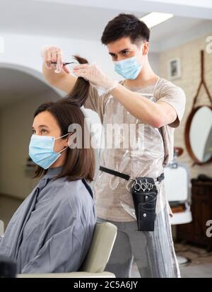 Positive männliche Friseur in Schutzmaske und Handschuhe machen Haar Styling für junge Frau, Arbeitstag im Schönheitssalon während Pandemie-Situation Stockfoto