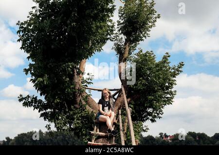 Eine junge Frau mit einem drei Monate alten schottischen Straight Kitten sitzt im Sommer auf einem schönen grünen Baum über dem See. Gehen, ruhen Sie mit einem Haustier. Die Blondine trägt kurze Denim-Shorts, ein schwarzes Baumwoll-T-Shirt und eine Brille. Stockfoto