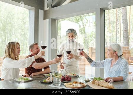 Älterer bärtiger Mann mit einem Glas Rotwein, der während des Abendessens mit Familienmitgliedern Toast trinkt Stockfoto