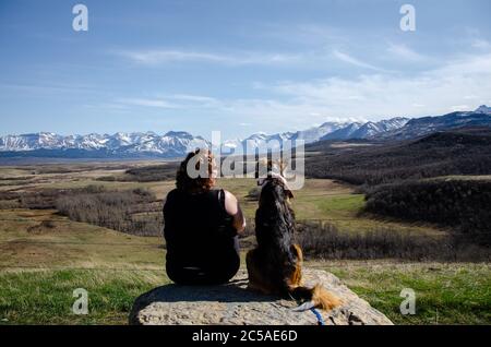 Frau mit ihrem Hund auf den Felsen sitzend und schauend An den schneebedeckten Bergen Stockfoto