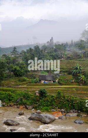 Wolkige Landschaft von Sapa, Vietnam. Die Berge verschwinden im Nebel. Felder und Bauernhöfe schmiegen sich an den Berg. Ein brauner Fluss im Vordergrund. Stockfoto