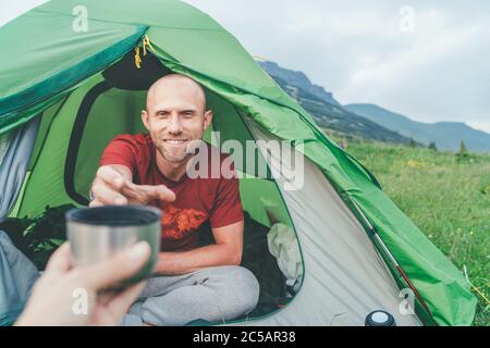 Lächelnder Glatze Mann im grünen Zelt, der die Thermoskanne für den morgendlichen Tee-Drink mit Berghintergrund nimmt. Aktive Menschen, die gerne reisen Stockfoto