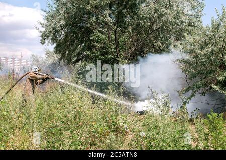 feuerwehrmann in einem Schutzanzug zwischen Gras und Bäumen löscht brennenden Müll in der Natur. Stockfoto