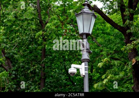 Eiserne Straßenlampe mit Videoüberwachungskamera im Park auf einem Hintergrund von grünen Bäumen. Stockfoto