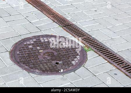 Ein rundes Mannloch und Gitter eines Entwässerungssystems auf einem Bereich aus quadratischen Steinfliesen, Nahaufnahme eines Regenwasserabflusssystems. Stockfoto