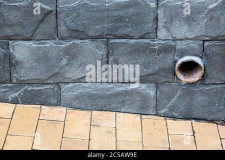 Eine Regenwasserleitung, die aus der dunkelgrauen Fliesenwand auf dem Gebäude in der Nähe mit braunen Steinfliesen aus nächster Nähe gepflastert. Stockfoto