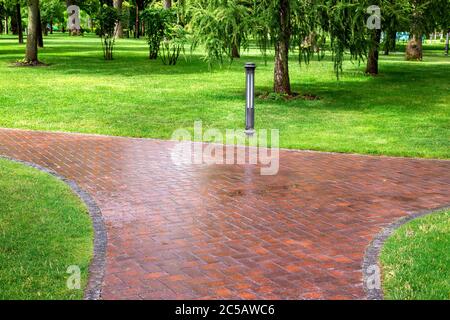 Nass nach Regen über einen Fußgängerweg in einem Park mit Grünflächen und Erdlaterne, leer niemand. Stockfoto