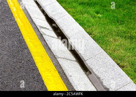 kanal ist ein Zementgraben eines Kanalisationssystems mit Regenwasserentwässerung auf der Seite einer Asphaltstraße mit gelben Markierungen und einem grünen Rasen. Stockfoto