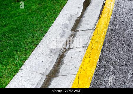 Fließendes Regenwasser im Kanal ist ein Zementgraben eines Drainagesystems auf der Seite einer Asphaltstraße mit gelben Markierungen und einem grünen Rasen. Stockfoto