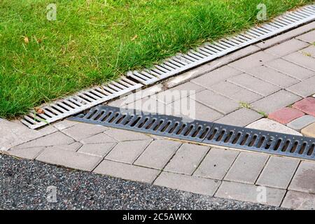 Die Kreuzung der Asphaltstraße und der Fußgängerweg aus Fliesen mit dem Gitter des Entwässerungssystems im Park mit grünem Gras gemacht. Stockfoto