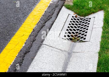 Kanalisationskantenwanne mit Betonrost für Regenwasserableitung in den Kanal auf asphaltierten Straße mit gelber Markierung und grünem Rasen. Stockfoto