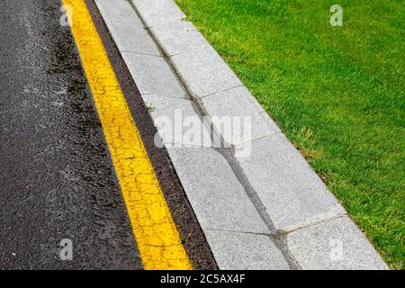 Regenwasser im Kanal ist ein Zementgraben eines Drainage-Systems auf der Seite der nassen Asphaltstraße mit gelber Linie und einem grünen Rasen auf Straßenrand Umwelt Stockfoto