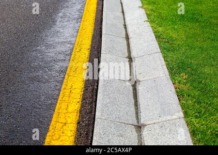 Fließendes Regenwasser im Kanal ist ein Zementgraben eines Drainage-Systems auf der Seite der nassen Asphaltstraße mit gelben Markierungen und einem grünen Rasen auf der Straße Stockfoto