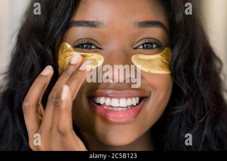 Anti-Aging, Hautpflege, Kosmetik-Konzept. Nahaufnahme Schönheit Porträt von charmanten lächelnden jungen afrikanischen Frau Anwendung goldenen medizinischen Augenflecken Stockfoto