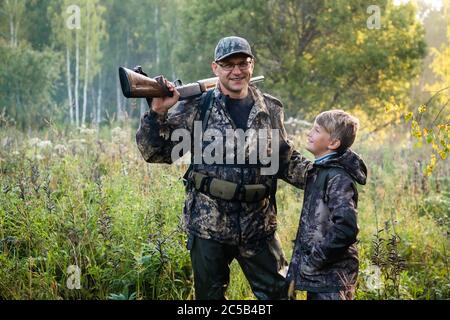 Der lächelnde Jäger oder Militärperson mit Gewehr oder Gewehr und sein Lehrling im Sommerwald während der Vogeljagd. Stockfoto