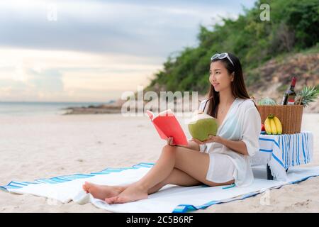 Junge asiatische Frau trägt Sonnenbrille ist mit dem Lesen eines Buches im weißen Sandstrand und in der Nähe des Meeres mit tropischen Früchten im Hintergrund entspannen. Sommer, holi Stockfoto