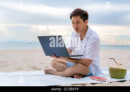 Junger asiatischer Mann, der während seiner Urlaubszeit mit einem Laptop draußen arbeitet, während er am schönen Strand sitzt. Sommer, Urlaub, Urlaub und glückliche Menschen ich Stockfoto