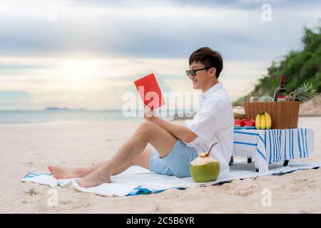 Junge asiatische Mann tragen Sonnenbrille ist mit einem Buch im weißen Sandstrand und in der Nähe des Meeres mit tropischen Früchten im Hintergrund entspannen. Sommer, Urlaub Stockfoto