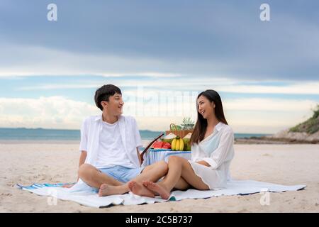 Asiatisches junges Paar sitzt auf der Picknickdecke und entspannen Sie sich am weißen Sandstrand und in der Nähe des Meeres mit tropischen Früchten im Hintergrund. Sommer, Urlaub, V Stockfoto