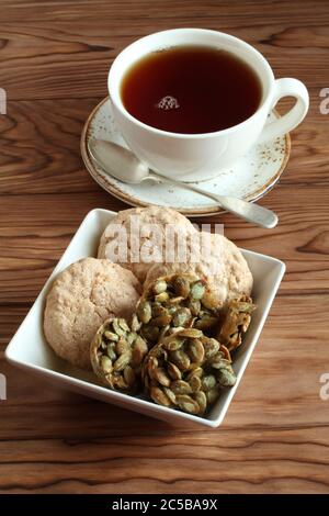 Mandelkekse und mit Zucker überzogene Kürbiskerne-Kekse in einer quadratischen Schüssel und eine Tasse schwarzen Tee auf einem Holztisch. Nahaufnahme Stockfoto