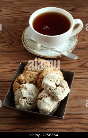 Haferflocken mit Walnüssen und Rosinen, Baiser mit Haselnüssen in einer quadratischen Schüssel und eine Tasse schwarzen Tee auf einem Holztisch. Nahaufnahme Stockfoto