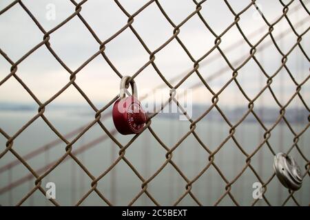 Liebesschlösser auf der Golden Gate Bridge, ein rotes Zahlenschloss, ein rundes Vorhängeschloss. San Francisco Skyline im Hintergrund. Grauer Himmel. Stockfoto