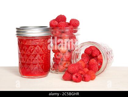 Ganze frische Himbeeren in offenen Maurer-Gläser, ein verschüttetes Obst auf dem Tisch vor voll hausgemachten Dosenkonfitüren auf hellem Holztisch, isol Stockfoto