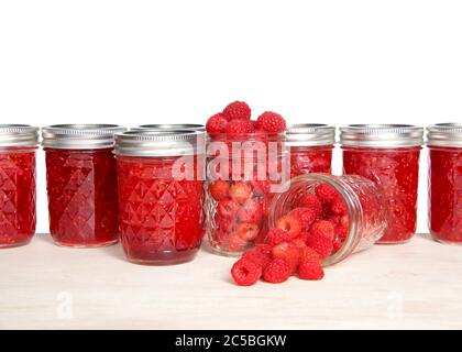 Ganze frische Himbeeren in offenen Maurer-Gläser, ein verschüttetes Obst auf dem Tisch vor voll hausgemachten Dosenkonfitüren auf hellem Holztisch, isol Stockfoto