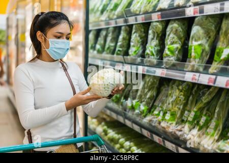 Junge asiatische Frau, die Gemüse beim Einkaufen von Lebensmitteln im Supermarkt und ihr Tragen einer medizinischen Maske zur Prävention Coronavirus (covid-19) Pandemie. n Stockfoto