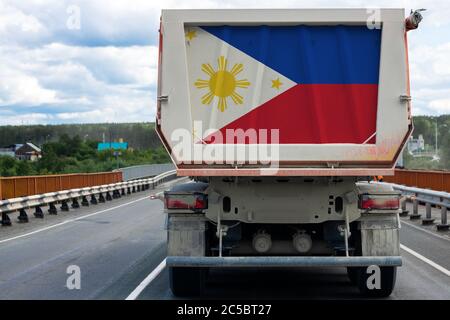 Großer LKW mit der Nationalflagge der Philippinen, der sich auf der Autobahn bewegt, vor dem Hintergrund der Dorf- und Waldlandschaft. Konzept des Exports Stockfoto