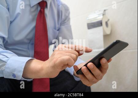 Geschäftsmann sitzt auf der Toilette im Badezimmer und mit einem Smartphone. Stockfoto