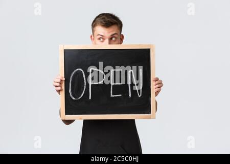 Kleineinzelhandelsinhaber, Café und Restaurant Mitarbeiter Konzept. Dumm schöner Barista, Verkäufer schaut weg, hält Schild in der Nähe des Gesichts offen Stockfoto