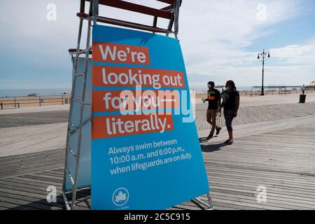 New York, USA. Juli 2020. Die Menschen gehen an einer Tafel über Badezeit an einem Strand in Coney Island in New York City, USA, 1. Juli 2020 vorbei. Am Mittwoch sind acht Strände in New York offiziell zum Schwimmen geöffnet, wenn Rettungsschwimmer täglich von 10 bis 18 Uhr im Einsatz sind.Quelle: Wang Ying/Xinhua/Alamy Live News Stockfoto