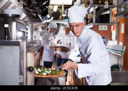 Jungen Koch in der Nähe von professionellen Backofen im Inneren des modernen Restaurant Küche, hält in seinen Händen Schaufel mit gekochtem Pizza Stockfoto